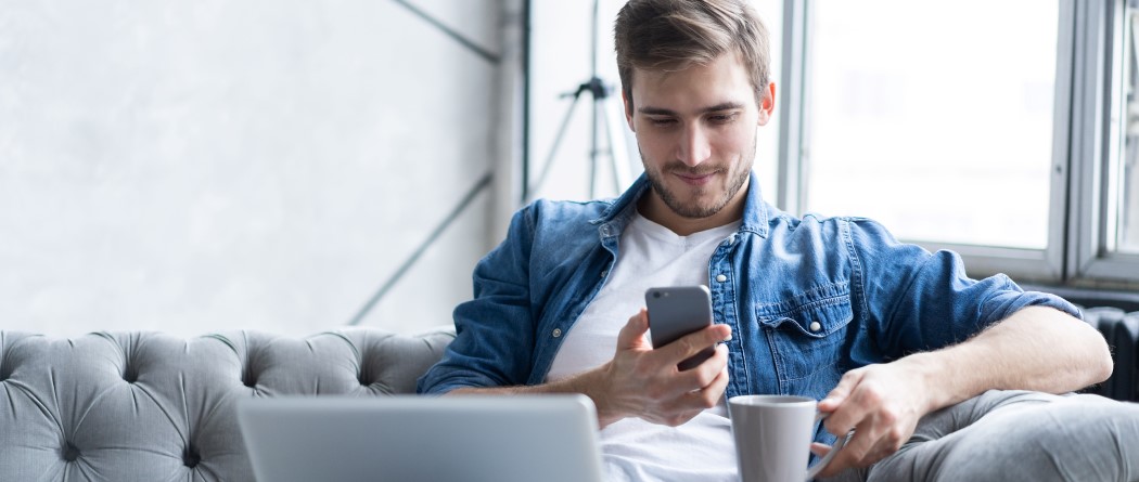 Man on couch drinking coffee, looking at phone with laptop on lap.