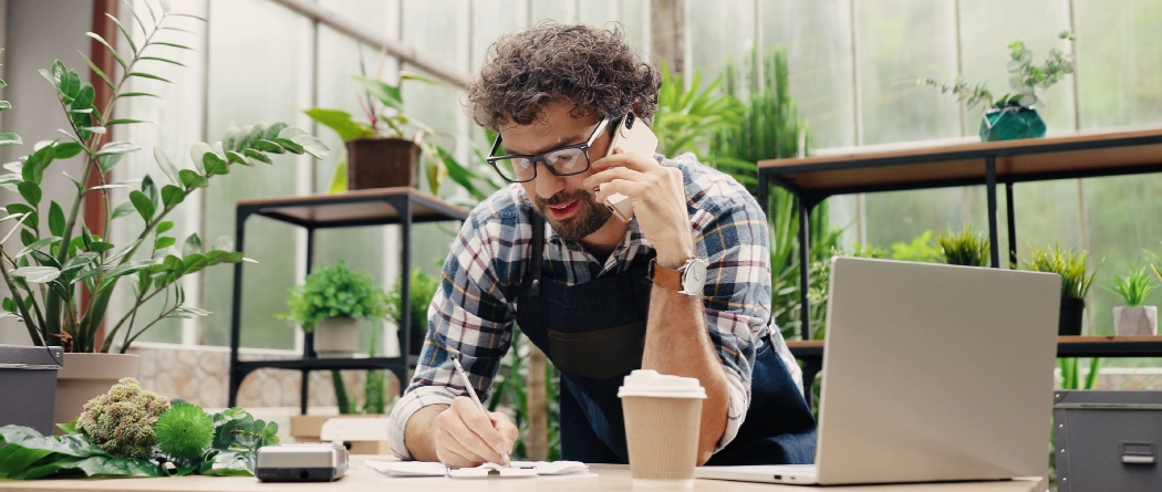 Man speaking on phone. Surrounded by plants and leaning over desk.