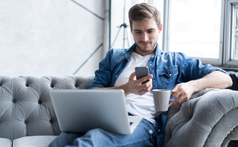 Man on couch drinking coffee, looking at phone with laptop on lap.